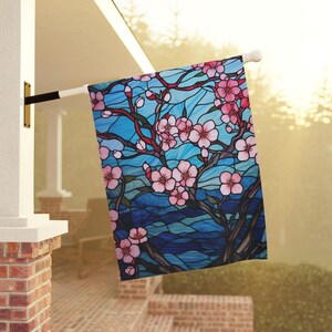 May include: A stained glass style garden flag with a blue background and pink cherry blossoms. The flag features a stylized tree with branches and leaves.