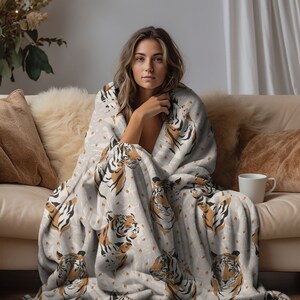 May include: A soft, plush throw blanket featuring a repeating pattern of tiger heads in shades of orange, black, and white against a light gray background. The blanket is draped over the shoulders, showcasing the detailed animal print design.