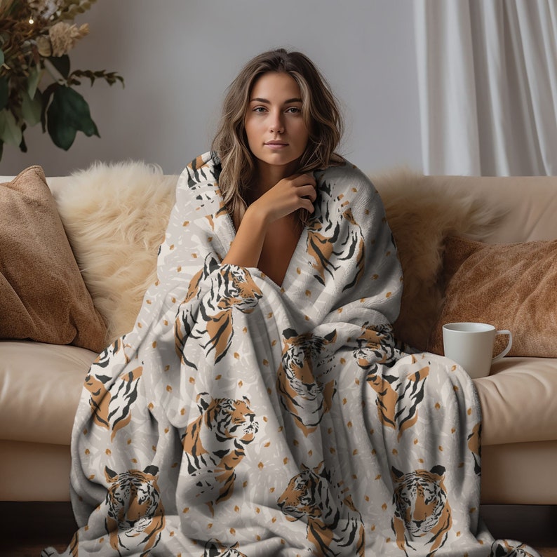 May include: A soft, plush throw blanket featuring a repeating pattern of tiger heads in shades of orange, black, and white against a light gray background. The blanket is draped over the shoulders, showcasing the detailed animal print design.