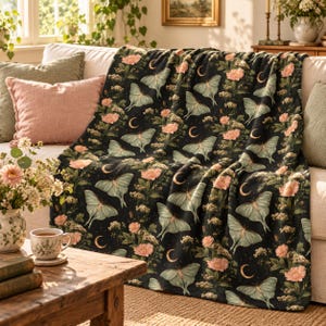 May include: A black throw blanket featuring a repeating pattern of pale green luna moths, soft pink peony flowers, and golden crescent moons. The fabric has a soft, plush texture and is draped over a sofa with decorative pillows.