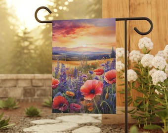 Puede incluir: Una bandera de jardín con una pintura vibrante de un campo de amapolas rojas, flores silvestres moradas y trigo dorado bajo un cielo de puesta de sol. La bandera se muestra en un poste de jardín de metal negro.