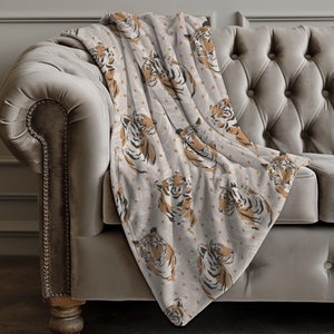 May include: A soft throw blanket featuring a repeating pattern of tiger heads in shades of orange, black, and white against a light gray background. The design displays multiple tiger faces oriented in various directions across the fabric surface.