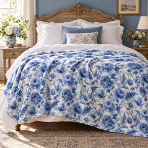 May include: A bed blanket featuring a blue and white floral pattern with detailed illustrations of peonies and ferns. The fabric is draped over a bed with white pillows and a blue accent pillow, showcasing a repeating botanical design.