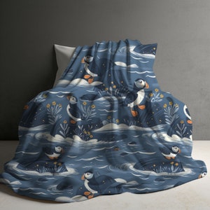 May include: A soft blanket featuring a repeating pattern of Atlantic puffins on a dark blue background. The design includes white waves, stylized rocks, and small yellow flowers interspersed among the birds, which have black and white feathers.