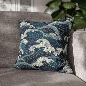 Puede incluir: Cojín decorativo cuadrado con un patrón estilizado de olas oceánicas en tonos azul marino, verde azulado y crema. El cojín está colocado sobre un sofá gris, mostrando su diseño y uso potencial como decoración del hogar.