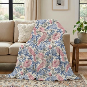 May include: A soft throw blanket featuring a repeating pattern of light pink birds, blooming pink roses, and intricate blue floral scrollwork on a cream-colored background. The fabric displays a detailed botanical design with soft textures.