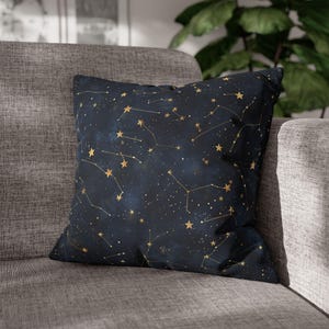 Peut inclure: Un coussin carré décoratif avec un fond bleu foncé orné d'un motif de constellations dorées. Le coussin est posé sur un canapé gris, avec une plante en arrière-plan. Le motif comprend des étoiles et des lignes dorées.