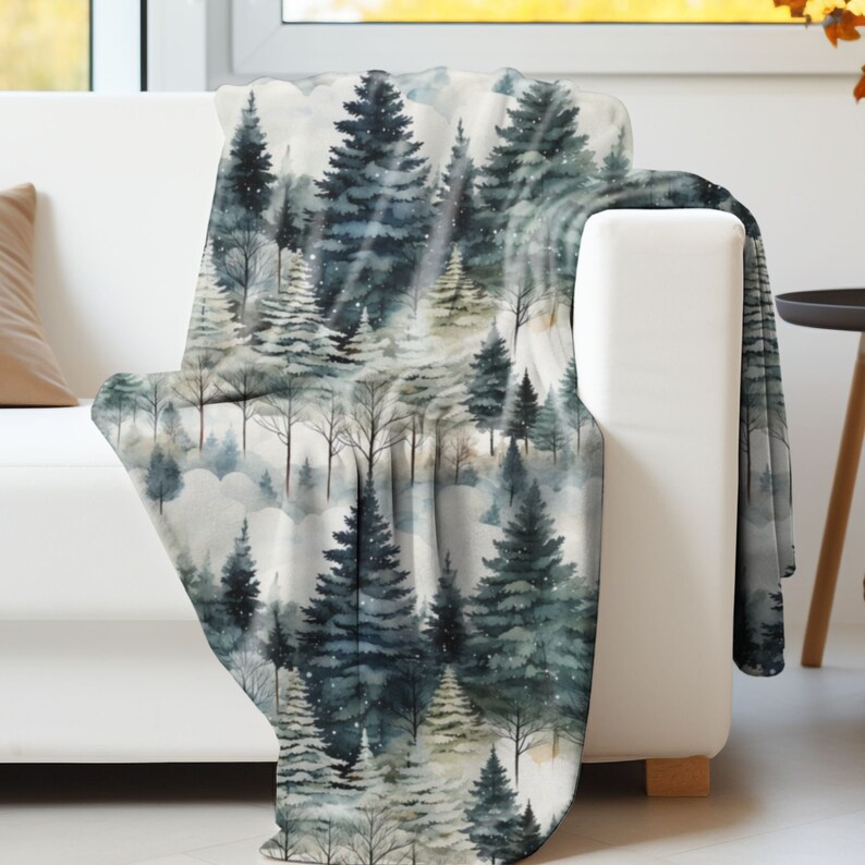 May include: A soft, plush throw blanket featuring a watercolour design of a snowy forest with tall, dark green pine trees. The background is a light grey with subtle hints of white and gold.