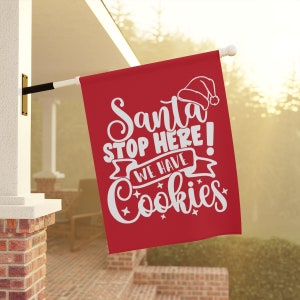 May include: Red Christmas flag with white text that reads "Santa Stop Here! We Have Cookies". The flag has a white Santa hat graphic at the top.