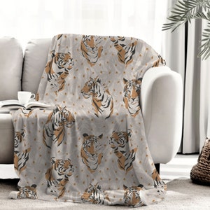 May include: A soft throw blanket featuring a repeating pattern of tiger heads in shades of orange, black, and white. The background is a light gray color with subtle, faint leaf-like shapes scattered throughout the fabric design.