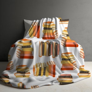 May include: A white throw blanket featuring a repeating pattern of stacked books in shades of orange, yellow, brown, and black. Some book spines are clearly printed with the text "JUST ONE MORE CHAPTER" in a simple, legible font style.