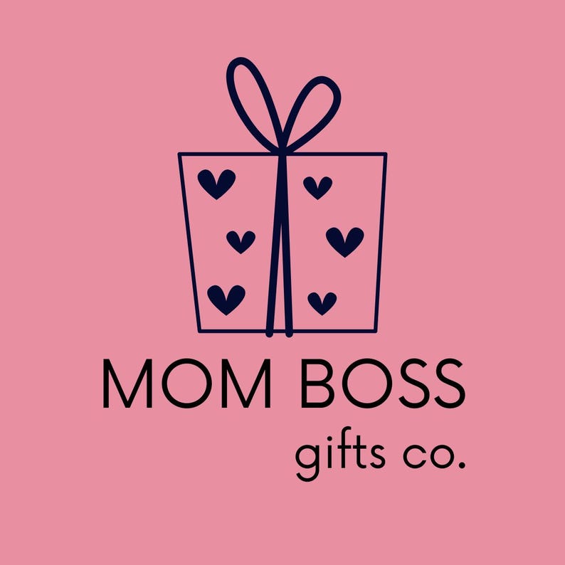 May include: A logo on a pink background featuring a dark blue line drawing of a square gift box tied with a bow and decorated with small hearts. Below the box, the text reads MOM BOSS gifts co. in a clean, dark blue sans-serif typeface.