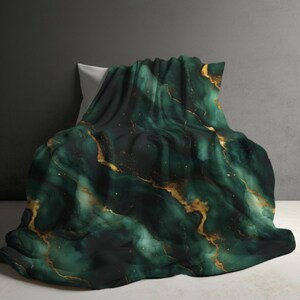 May include: A plush blanket with a dark emerald green marble design and gold veining. The blanket is draped over a white pillow, creating a luxurious and elegant aesthetic. The blanket's design resembles natural stone.