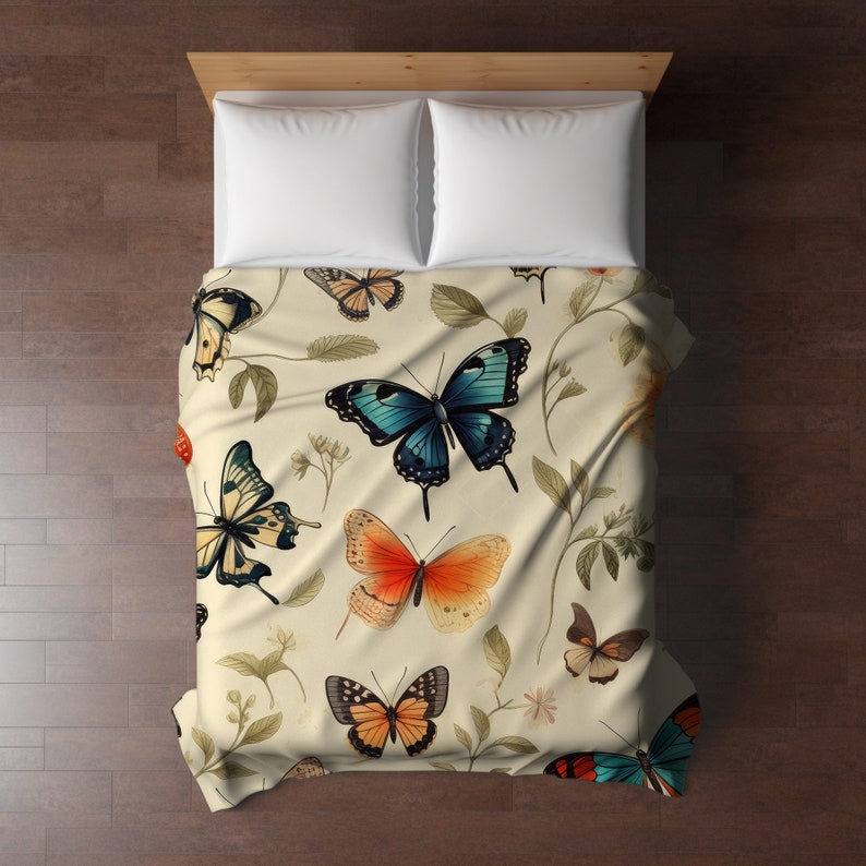 Op de afbeelding: Een cr&egrave;mekleurige sprei met een herhalend patroon van diverse vlinders in tinten blauw, oranje en zwart, afgewisseld met delicate botanische illustraties van dunne stengels en groene bladeren, die een bed met twee witte kussens bedekt.