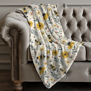May include: A white fleece throw blanket with a pattern of yellow flowers, green leaves, and black and yellow bees.