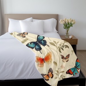 Op de afbeelding: Een cr&egrave;mekleurige plaid met een herhalend patroon van diverse vlinders in tinten blauw, oranje en bruin, afgewisseld met delicate botanische illustraties van groene bladeren en kleine steeltjes over het oppervlak van de stof.