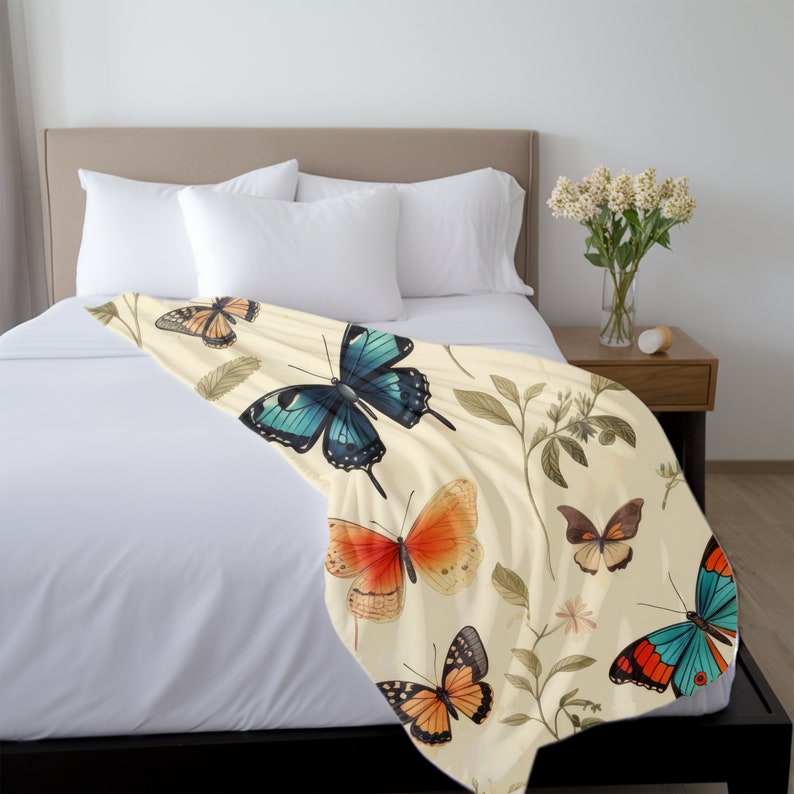 Op de afbeelding: Een cr&egrave;mekleurige plaid met een herhalend patroon van diverse vlinders in tinten blauw, oranje en bruin, afgewisseld met delicate botanische illustraties van groene bladeren en kleine steeltjes over het oppervlak van de stof.