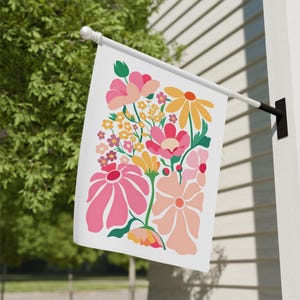 Puede incluir: Una bandera blanca para exteriores con un diseño floral colorido. El diseño incluye flores rosas, amarillas y melocotón con hojas verdes. La bandera está montada en un poste blanco y se muestra fuera de un edificio.