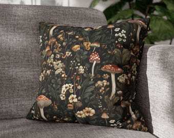 May include: Square decorative pillow with a dark background featuring a botanical print of mushrooms, flowers, and foliage in shades of red, white, brown, and green. The pillow is placed on a gray sofa.
