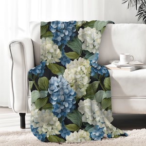 May include: A rectangular throw blanket featuring a repeating botanical pattern of blue and white hydrangea flower clusters interspersed with green leaves against a solid black background. The fabric displays a soft texture with a floral print.