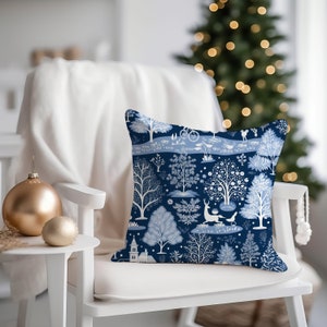 K&ouml;nnte beinhalten: Ein blaues und wei&szlig;es Kissen mit einer winterlichen Waldszene mit B&auml;umen, V&ouml;geln, einem Reh und einem Fahrrad. Das Kissen liegt auf einem wei&szlig;en Stuhl vor einem Weihnachtsbaum.