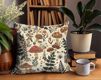 Op de afbeelding: Vierkantig sierkussen met een crèmekleurige achtergrond met een patroon van paddenstoelen, varens en wilde bloemen in bruin-, rood- en groentinten. Het kussen ligt op een houten oppervlak met planten en boeken op de achtergrond.