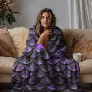 May include: A soft throw blanket featuring a repeating pattern of overlapping dark purple and black scales. The design mimics a textured dragon or mermaid scale aesthetic with deep violet highlights and subtle cracked details across the surface.