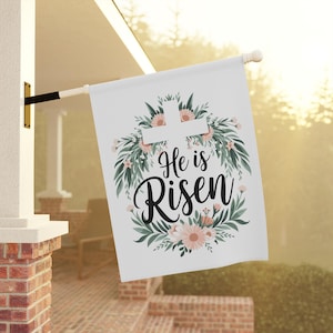 以下が含まれることがあります： 白いガーデンの旗で、花輪のデザインが施されています。花輪にはピンクの花、緑の葉、白い十字架が飾られています。"He is Risen"という文字が黒い筆記体で書かれています。