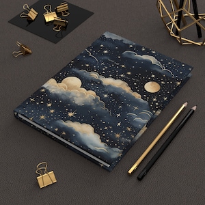 May include: A hardback journal with a celestial design. It features a navy blue background, gold stars, and moon illustrations. Gold and black pencils and gold binder clips are also present.