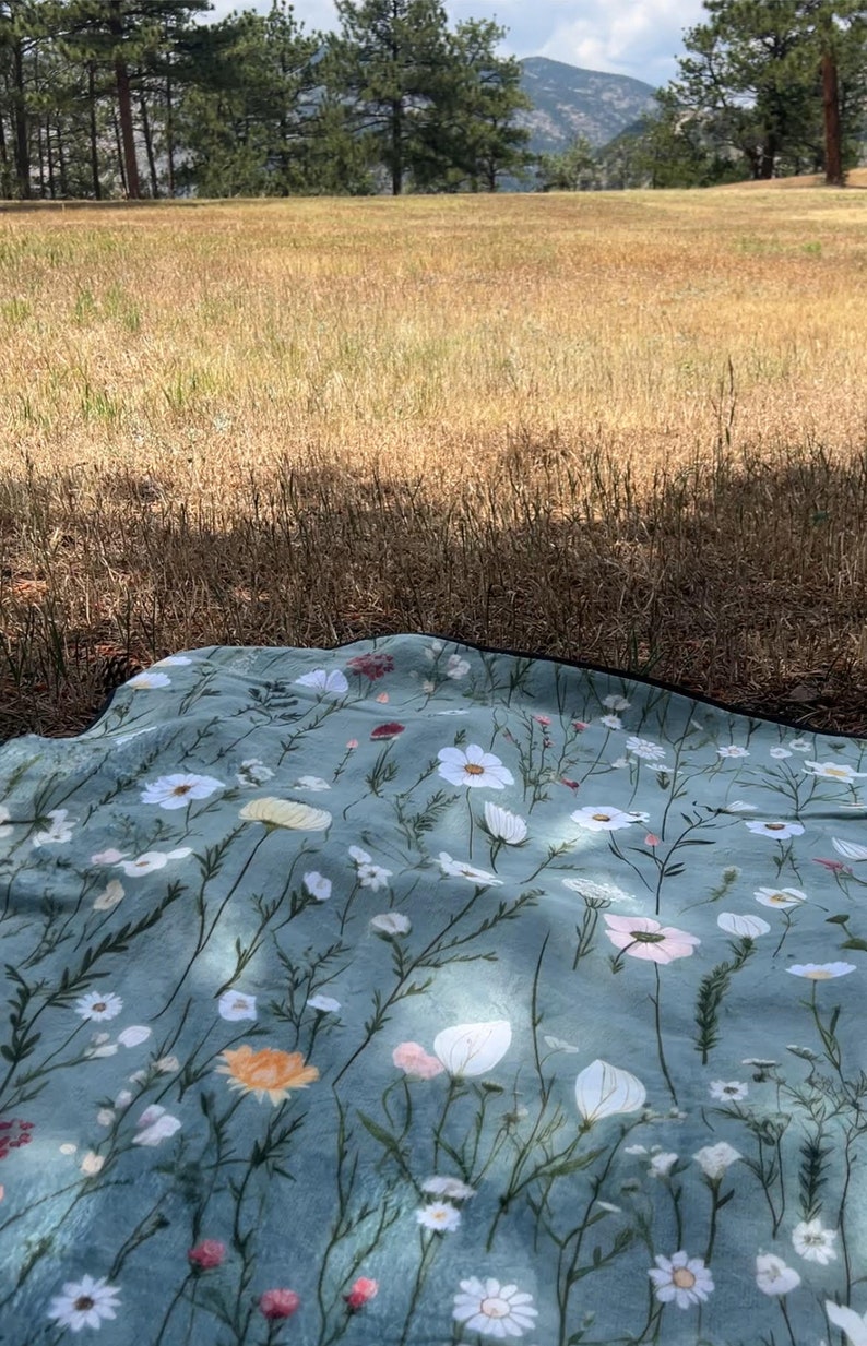 May include: A blue picnic blanket with a floral pattern featuring white, pink, and yellow flowers. The blanket is laid out on a grassy field with trees in the background.