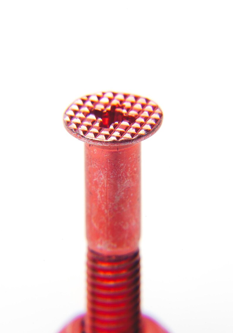 以下が含まれることがあります： テクスチャ加工された円形の頭部を持つ赤いボルトのクローズアップ。頭部は格子状のパターンと中央に小さな穴があります。ボルトのシャフトは円筒形でねじ切りされており、鮮やかな赤色です。