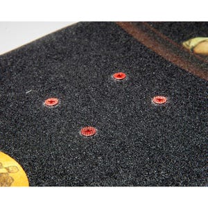 以下が含まれることがあります： 黒色のテクスチャード加工された表面を持つスケートボードデッキのクローズアップ。4つの赤い円形のハードウェアパーツが見えます。デッキには茶色の縁と、隅に黄褐色と茶色のデザインがあります。