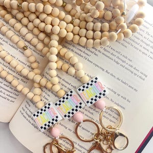 Peut inclure: Un porte-clés avec un charme à carreaux rose et arc-en-ciel avec le mot "Teacher" dessus. Le charme est attaché à un anneau en métal doré et une chaîne en métal doré. Le porte-clés est posé sur un livre avec du texte.