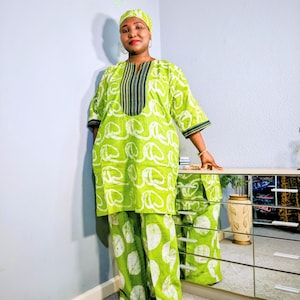 Op de afbeelding: Een limoengroene tweedelige outfit met een bijpassende hoofddoek. De tuniek heeft een wit wervelpatroon en een donkergroene en zwart gestreepte halslijn. De broek met wijde pijpen heeft een wit cirkelvormig patroon. De outfit is gemaakt van een lichte stof.