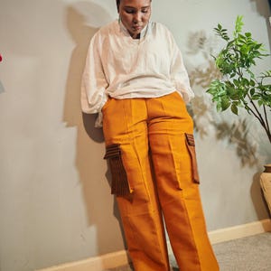 Peut inclure: Une personne porte un chemisier blanc, un pantalon cargo orange et un couvre-chef violet à motifs. Le pantalon a deux poches rayées marron et noir. La personne est appuyée contre un mur gris.