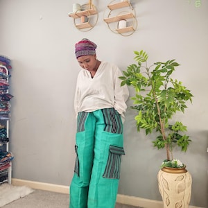 May include: A person wearing a white long-sleeve shirt and teal cargo pants with black and green striped accents. The pants have large pockets on the sides. A patterned head wrap is also visible.