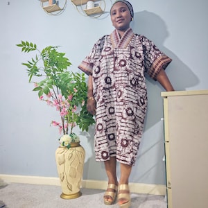May include: A knee-length dress with a brown and white pattern, short sleeves, and a V-neck. The dress is worn with gold platform sandals. A black head covering is also worn. The background features a vase with flowers and wall shelves.