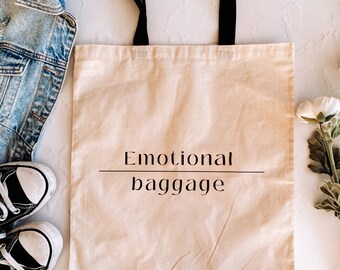 May include: A beige canvas tote bag with black handles and the text "Emotional baggage" printed in black.