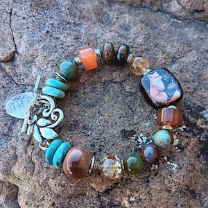 May include: A bracelet with a silver clasp and a variety of colorful beads, including turquoise, brown, orange, and green. The bracelet also features a square bead with a picture of a fox.