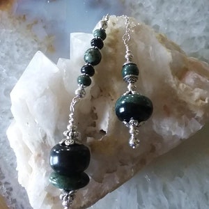 May include: Two green and black gemstone pendulums with silver chains and beads. The pendulums are hanging from a white and brown agate geode.