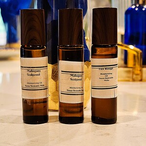 May include: Three small, brown glass bottles with dark wooden caps. Each bottle has a white label with black text. The labels read "Mahogany Teakwood" and "Lux Rouge". The bottles contain moisturizing oil.