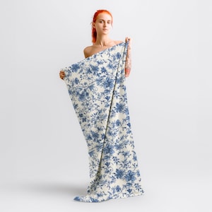 May include: A woman is holding a white fabric with a blue floral pattern. The fabric is draped over her body and has a vintage style.