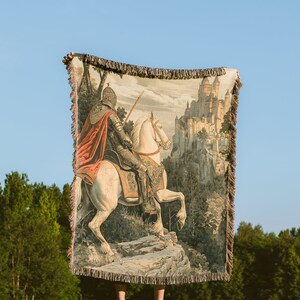 May include: A tapestry woven with a knight on horseback riding towards a castle in the distance. The knight is wearing armor and a red cape. The horse is white and has a brown saddle. The castle is made of stone and has many towers. The tapestry is brown and beige with a fringe border.