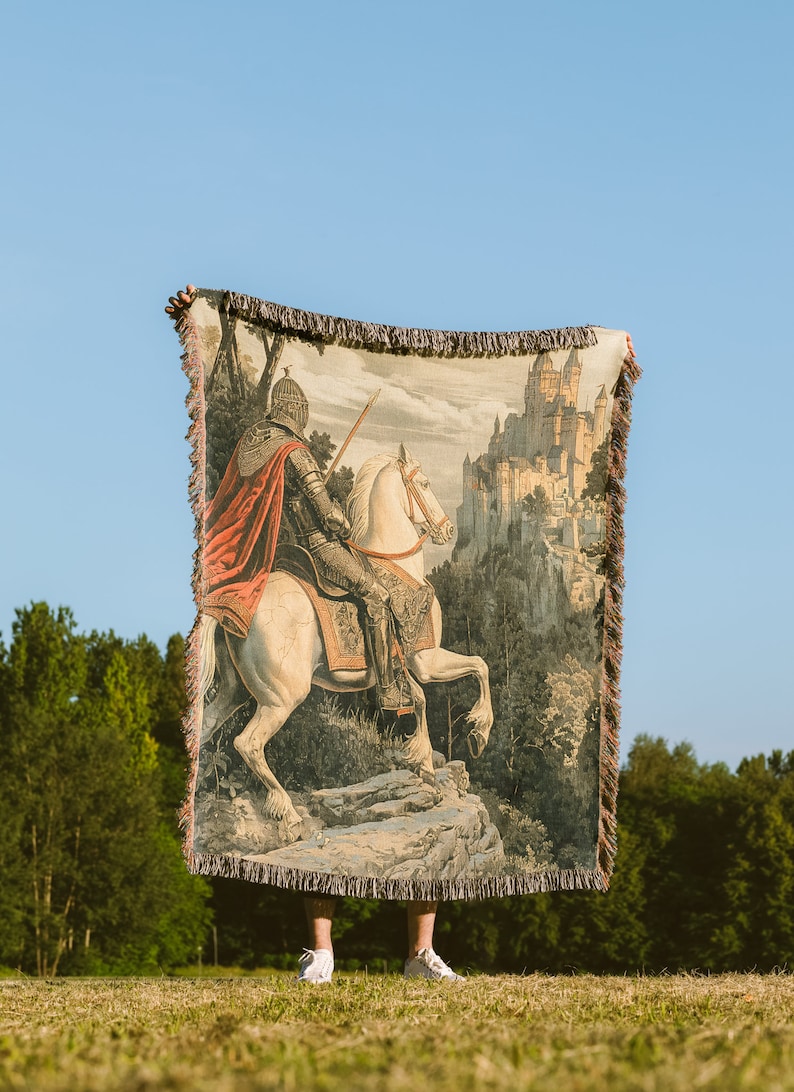 May include: A tapestry woven with a knight on horseback riding towards a castle in the distance. The knight is wearing armor and a red cape. The horse is white and has a brown saddle. The castle is made of stone and has many towers. The tapestry is brown and beige with a fringe border.