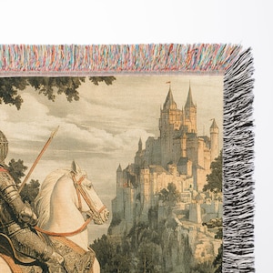 May include: A tapestry featuring a knight on horseback riding towards a large, stone castle. The knight is wearing armor and a red cape. The castle is perched on a hilltop and has many towers. The tapestry has a fringed border.