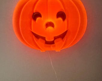 May include: A bright orange, glowing plastic pumpkin wall decoration featuring a carved jack-o'-lantern face with two crescent-shaped eyes, a small nose, and a wide, toothy grin. A thin white power cord hangs down from the base to a wall outlet.