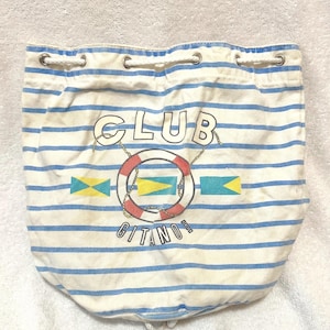 May include: A white drawstring bag featuring horizontal blue stripes. The front displays the text CLUB GITANO in black lettering, centered around a red and white life preserver graphic with three small yellow and teal nautical flag icons.