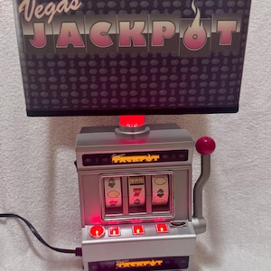 May include: A table lamp featuring a rectangular shade with a dark gray background, a repeating dollar sign pattern, and the text Vegas JACKPOT. The base is a silver-colored slot machine replica with three spinning reels, a red lever, and red lights.