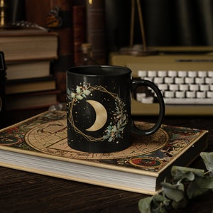 Könnte beinhalten: Eine schwarze Keramiktasse mit einem goldenen Halbmond und einem floralen Design auf dunklem Hintergrund. Die Tasse steht auf einem alten Buch mit einer Weltkarte auf dem Einband.