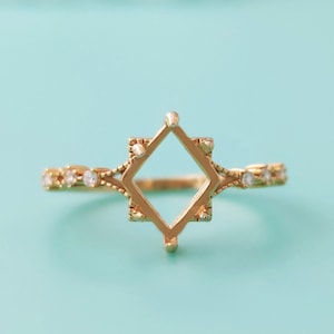 May include: A gold-colored ring with a square-shaped center setting. The band features small, clear gemstones and decorative detailing. The ring is set against a light blue background, showcasing its intricate design and craftsmanship.